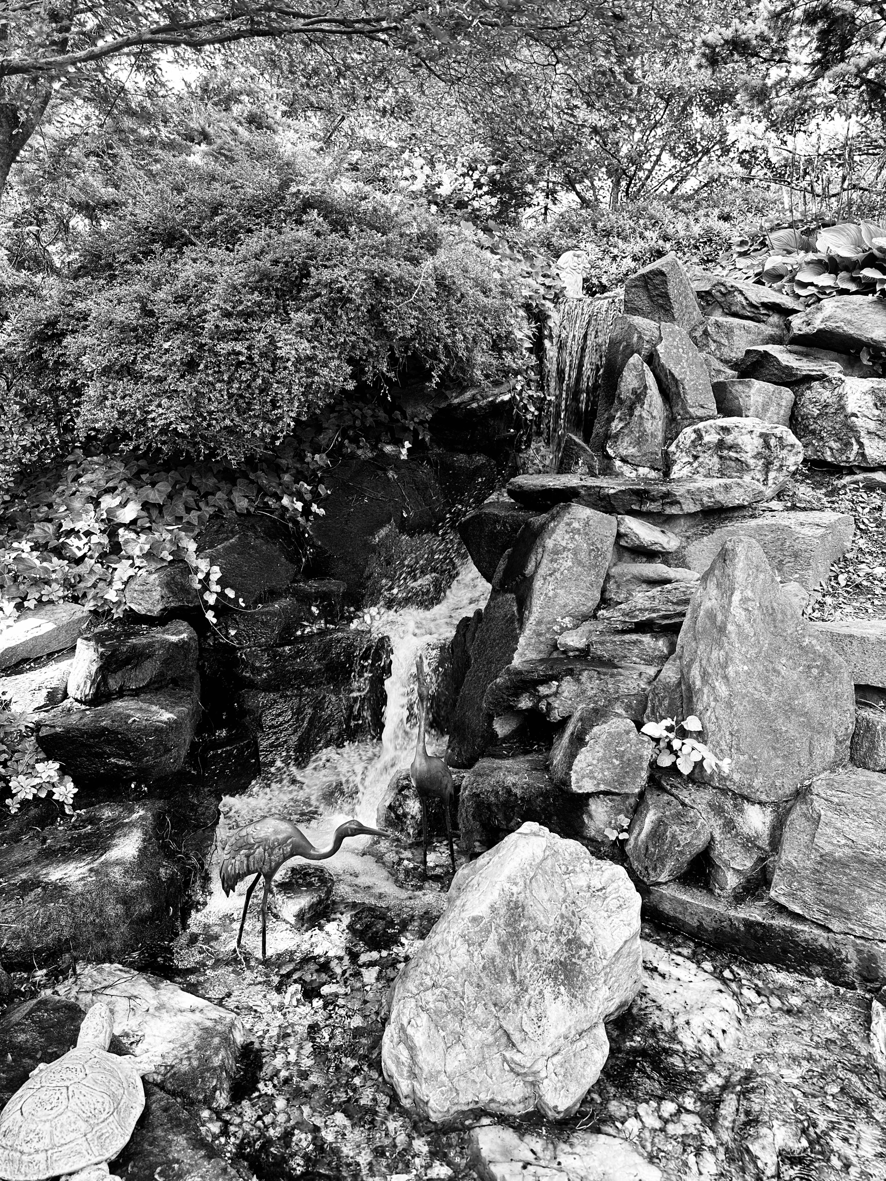 Black and white photo of a small stone waterfall in a Japanese-style garden with a metal statue of an ibis at the base.