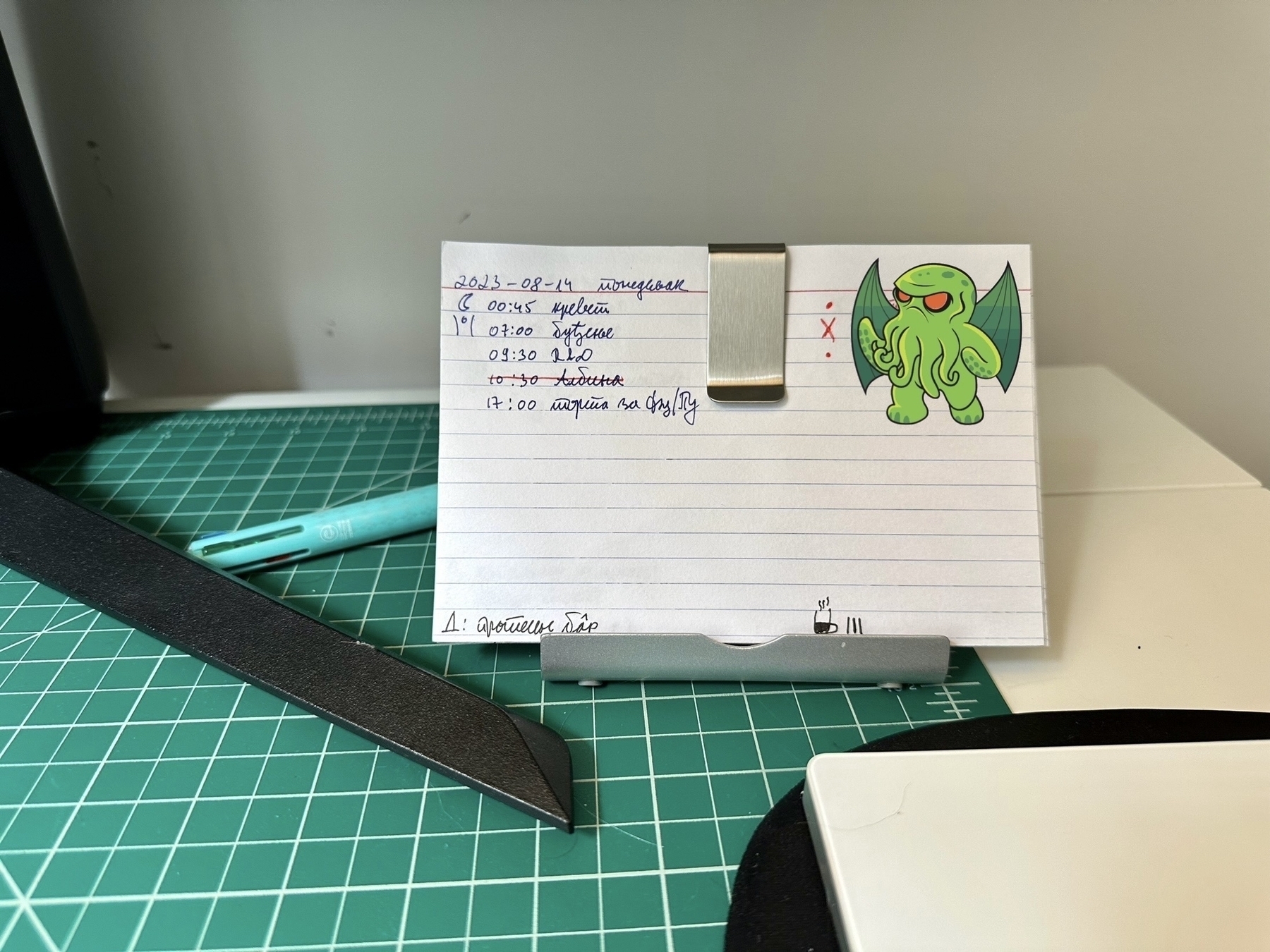 Close-up photo of a stack of index cards propped up on a phone stand, held by a metal clip. The front card has a task list and calendar for August 14, 2023. Most of it is in unreadable Cyrillic cursive.